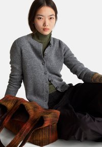 Šedý kardigan s knoflíkovým zapínáním, přeložený přes zelený rolák. Vedle ní je texturovaná, vícebarevná plstěná taška v oranžové a hnědé barvě.