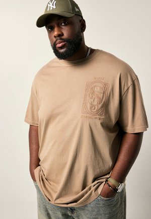 Beige bomulds-t-shirt med et præget logodesign på venstre bryst, parret med lysvasket denim. Accessories inkluderer en grøn kasket.