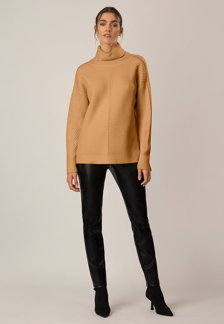 Beige gerippter Strickpullover mit Stehkragen in lässigem Schnitt, kombiniert mit schwarzen Kunstlederhosen und schwarzen Ankle Boots.