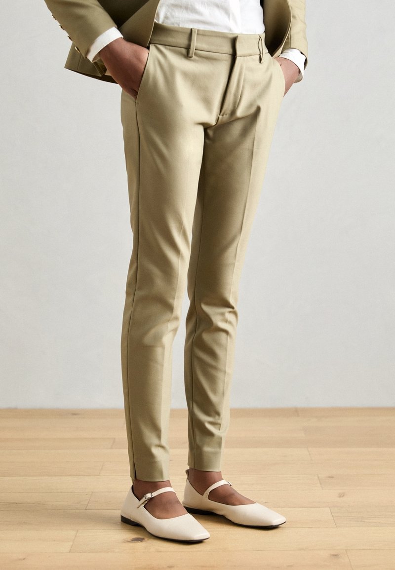 Femme portant un pantalon beige ajusté avec les mains dans les poches et des chaussures plates blanches Mary Jane, debout sur un sol en bois.
