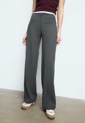Persoon draagt donkergrijze hoge taille wijde broek, donker mouwloos topje en beige-witte sneakers, staand op een betonnen vloer.