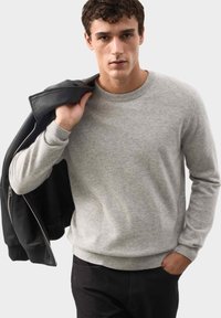 Jasnoszara sweter wykonany z miękkiego, teksturowanego materiału, z okrągłym dekoltem i prążkowanymi mankietami. Model trzyma czarną kurtkę na ramieniu.