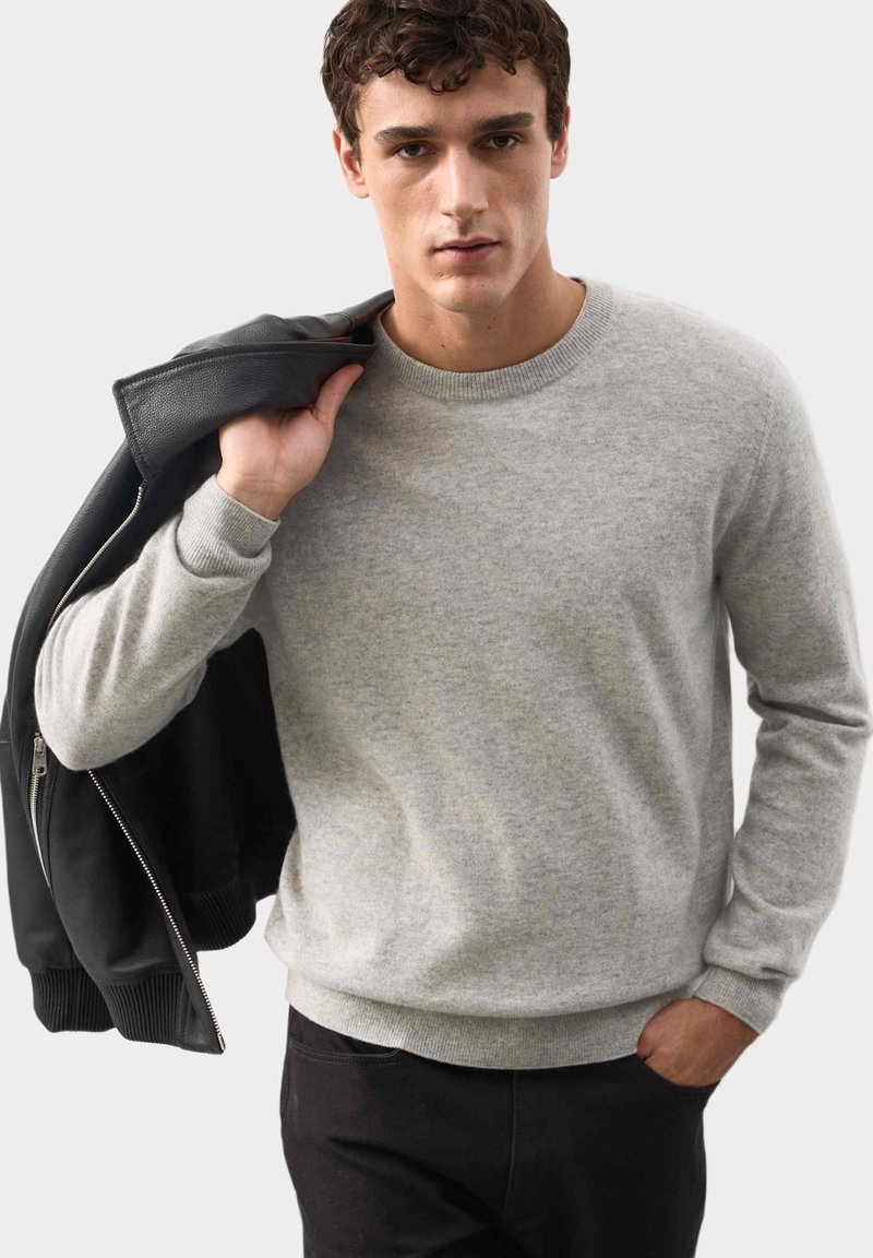 Jasnoszara sweter wykonany z miękkiego, teksturowanego materiału, z okrągłym dekoltem i prążkowanymi mankietami. Model trzyma czarną kurtkę na ramieniu.