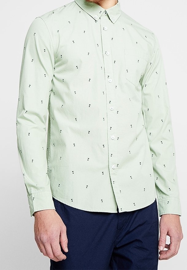 Chemise vert clair à manches longues avec boutons, présentant un petit motif noir abstrait, portée avec un pantalon bleu marine foncé.