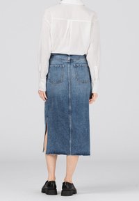 Kvinna iklädd en vit långärmad blus instoppad i en hög midja, knälång jeanskjol med bakfickor och en sidofick.