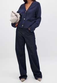 Granatowy dzianinowy kardigan z dekoltem w serek, ściągaczami na mankietach i złotymi guzikami, zestawiony z luźnymi ciemnymi jeansami i jasnoszarą torebką.