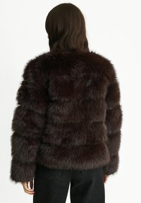 Brown faux fur jacket with a textured, striped design, featuring long sleeves and a high collar. Worn over black pants.