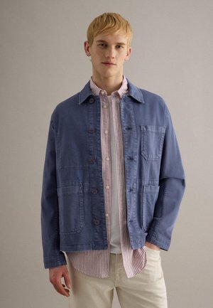 Junger Mann mit kurzem blondem Haar, der eine blaue Jacke mit Knöpfen über einem rot-weiß gestreiften Hemd und hellbeigen Hosen trägt.