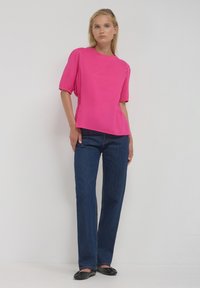 Donna in piedi contro un muro bianco, che indossa una blusa rosa brillante a maniche corte, jeans blu scuro e scarpe nere flat.
