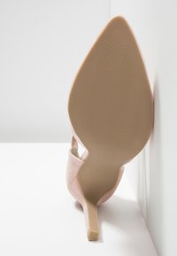 Chaussure à talon haut beige à bout pointu inclinée vers le haut, montrant la semelle texturée et le talon fin sur fond blanc.