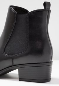 Stivale alla caviglia in pelle nera con pannello laterale elastico, linguetta sul retro e tacco basso impilato su sfondo bianco.