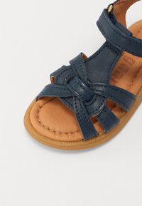 Marineblå lærsandaler med flettede stropper, tan fotseng og gummisåle. Har en gullfarget detalj på ankelstroppen.