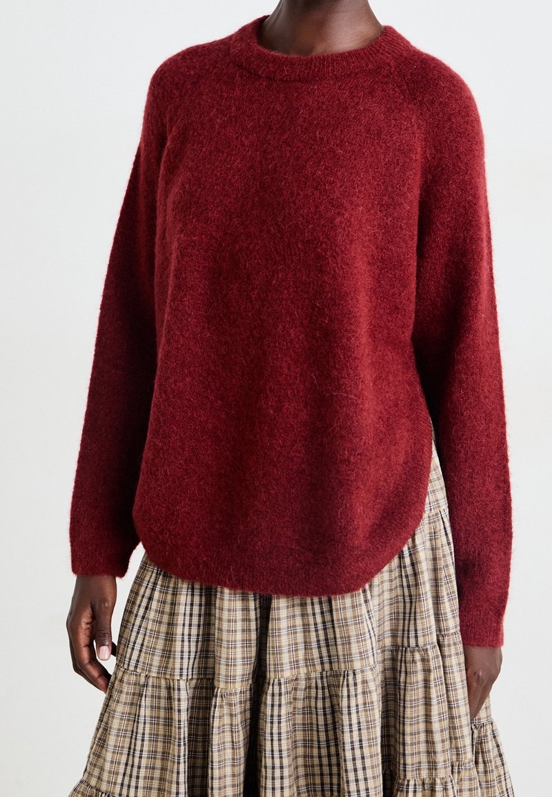Personne portant un pull rouge foncé à manches longues et duveteux associé à une jupe à volants beige et marron à carreaux sur fond blanc.