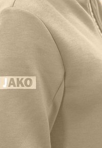 Felpa color beige chiaro con una superficie liscia e spalle arrotondate. Presenta un'etichetta rettangolare bianca con la scritta "JAKO" in lettere grigie.