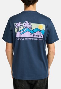 Donkerblauwe t-shirt met een grafisch ontwerp van palmbomen, bergen en zon in pasteltinten met het RVCA-logo en geografische coördinaten.