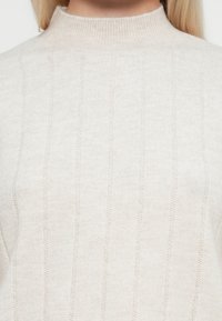 Vaalean beige neulepusero, jossa on korkea kaulus, valmistettu pehmeästä kankaasta. Siinä on pystysuuntainen ribbitekstuuri ja istuva siluetti.