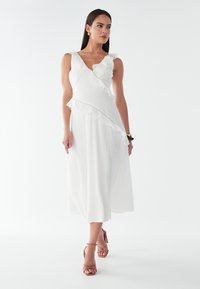 Balta midi sukienė su banguoto apykaklės ir šoninių akcentų, pasižyminti tekstūruota medžiaga ir laisva siluetu, derinama su dirželių sandalu.