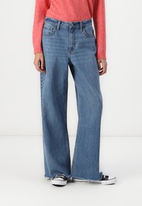 Jeans large de couleur bleu clair en denim, avec un ourlet effiloché, des poches avant et une taille haute, associés à des chaussures en toile noires.