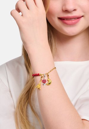 Gold charm bracelet featuring red beads, Eiffel Tower, heart-shaped, silver gymnast, and gold croissant charms, displayed on a wrist.