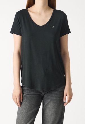 Kobieta ubrana w czarny T-shirt z krótkim rękawem i dekoltem w serek z małym, białym logo ptaka, w połączeniu z ciemnoszarymi dżinsami, stojąca na białym tle.