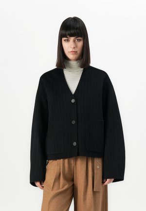 Vrouw in een zwarte knoopcardigan over een gestreepte coltrui en een bruine hoge taille wijde broek met riemdetails, staand tegen een witte achtergrond.