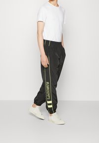 Svarta joggingsbyxor med neongula sidostråkar och "LACOSTE"-branding. Matchas med en enkel vit t-shirt och vita sneakers.