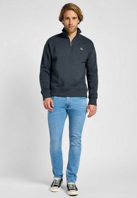 Felpa blu scuro con zip e colletto alto, polsini e vita a coste, abbinata a jeans azzurri e scarpe da ginnastica bianco e nero.