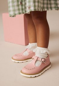 Chaussures roses en cuir à tige en T avec détails perforés, semelles blanches et boucle décorative, associées à des chaussettes blanches bordées de dentelle.