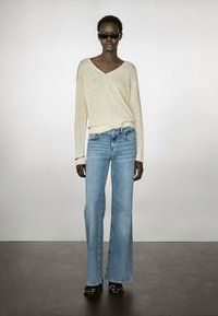 Maglione giallo chiaro a scollo a V in morbido maglificio; abbinato a jeans a vita alta e gamba larga blu. Il modello indossa occhiali da sole scuri e sandali con plateau.
