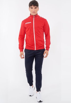 Mann trägt eine rote Jacke mit durchgehendem Reißverschluss und "LEGEA"-Logo, marineblaue Jogginghose und weiße Sneaker, steht vor einem schlichten weißen Hintergrund.