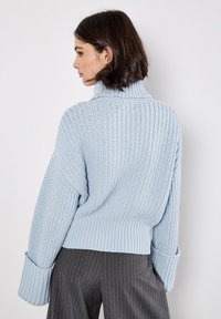 Maglione lavorato a maglia azzurro chiaro con colletto alto, texture a costine, maniche larghe e vestibilità ampia, abbinato a pantaloni a righe verticali grigio scuro.