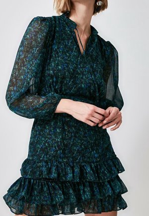 Femme portant une robe transparente fleurie vert foncé et bleue avec de longues manches bouffantes et un ourlet à volants superposés.