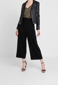 Veste de motard en cuir noir, blouse vert olive avec des détails de boutons, pantalons noir à jambes larges et sandales à talons noires. Fond neutre.