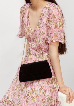 Femme portant une robe florale rose et marron avec des manches volantées, tenant une pochette en velours noir avec une chaîne dorée.