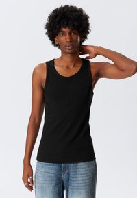 Crna rebrasta majica bez rukava s širokim naramenicama i okruglim izrezom, u kombinaciji sa svijetloplavim trapericama. Ton kože je srednji.