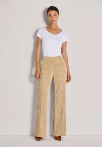 Beige Cordhosen im weiten Schnitt mit strukturiertem Stoff und seitlichen Taschen. Kombiniert mit einem taillierten weißen T-Shirt mit kurzen Ärmeln und braunen Absatzschuhen.