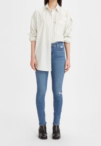 Camisa de botones blanca a rayas con mangas enrolladas, combinada con pantalones ajustados azules con detalles rasgados y botas negras hasta el tobillo.