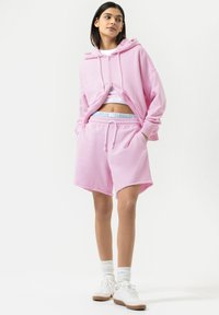 Roze cropped hoodie en bijpassende shorts gemaakt van zacht materiaal. De hoodie heeft een rits aan de voorkant, witte trekkoordjes en een losse pasvorm. Witte sneakers.