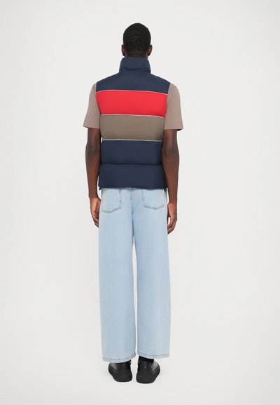 Kleurblok puffer vest met marineblauwe, rode en bruine secties; gecombineerd met wijde lichtblauwe denim en zwarte sneakers.