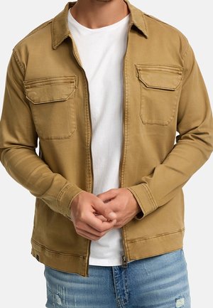 Mand iført en ubrugt tanjakke med klaplommer over en hvid t-shirt og slidte blå jeans, hænderne foldet ved taljen.