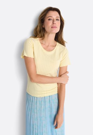 Hullámos hajú nő rövid ujjú, sárga bordás felsőt és világoskék redős szoknyát visel, fehér háttér előtt áll.