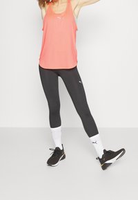 Koralowy sportowy top na ramiączkach w zestawieniu z czarnymi legginsami. Wykonany z lekkiego materiału z wyraźnymi nadrukami logo. Czarne buty sportowe i białe skarpety.