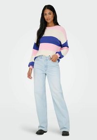 Femme portant un pull à rayures rose, crème et bleu, un jean à jambes larges bleu clair et des bottes noires, debout avec une main dans la poche.