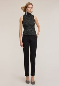 Czarny bezrękawnik z fakturowanej tkaniny i wysokim kołnierzem, w połączeniu z wąskimi czarnymi spodniami i szarymi szpilkami w szpic.