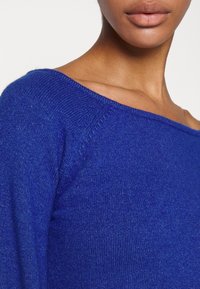 Modrý pletený svetr se širokým výstřihem typu bateau, s jemnou strukturou a žebrovanými detaily na výstřihu a ramenou.