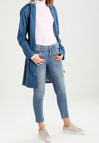 Blå vattentät regnjacka med tryckknappar, lagd över en vit polotröja. Buren med ljusblå skinny jeans och beige sneakers.