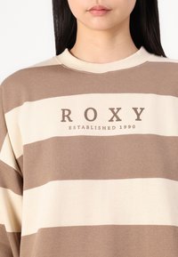 Stripet genser i myk stoff med vekslende brune og kremfargede striper, med "ROXY" og "ETABLERT 1990" trykt i mørkebrun.