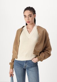 Veste bomber en daim marron avec des boutons à pression, portée sur un pull en maille écru à col en V. Jean bleu avec une silhouette ajustée et un sac bleu clair.
