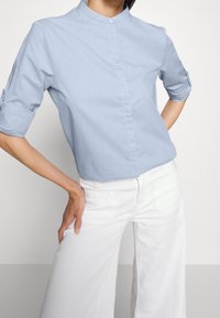 Person som bär en ljusblå knäppt skjorta med uppvikta ärmar och vita vida byxor, med handen vilande på höften mot en enfärgad bakgrund.