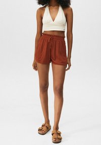 Oberteil mit Neckholder-Ausschnitt aus cremefarbenem Stoff, kombiniert mit rostfarbenen, strukturierten Shorts mit elastischem Bund. Braune Sandalen mit Schnallen vervollständigen den Look.